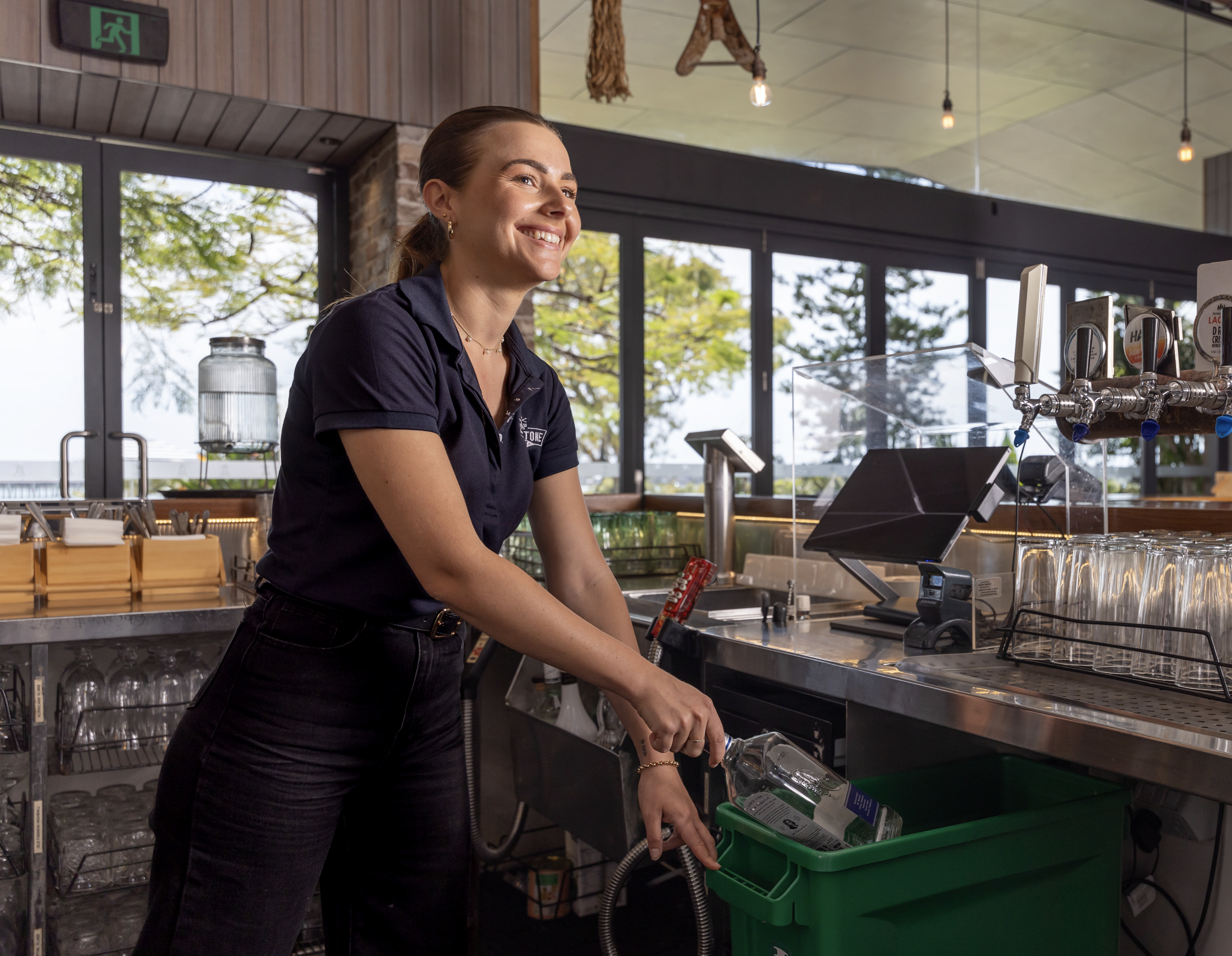 A hospitality worker behind a bar using a green bin for drink container collection.